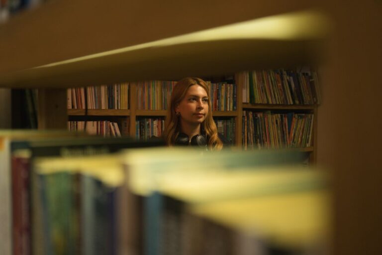 Young woman in a library looking thoughtfully through book shelves.
