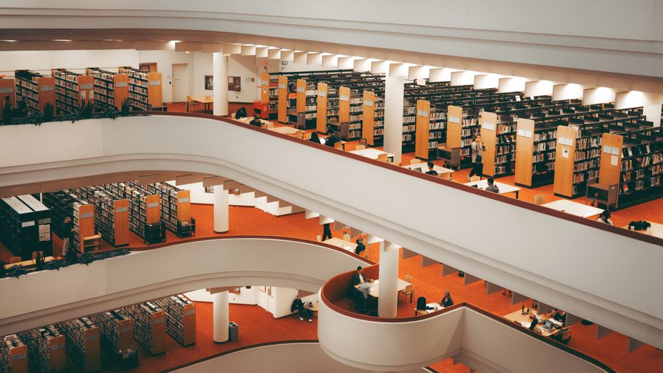 Three students reading on a staircase near bookshelves inside a library setting.