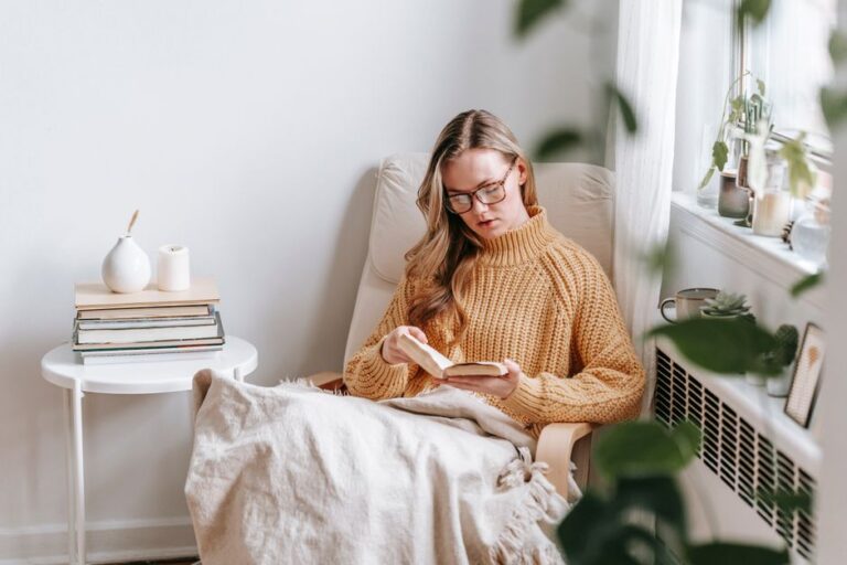 Young female student in trendy sweater and glasses sitting in white armchair with plaid near table with books and candle while reading book